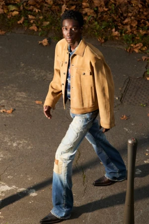 A young adult male in a tan jacket and distressed jeans is walking on a city street with leaves in the background during daytime.