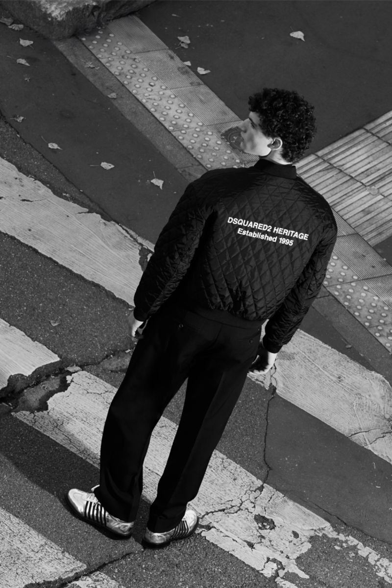 Black and white shot of a slender man wearing a quilted bomber jacket standing on a crosswalk with golden sneakers.