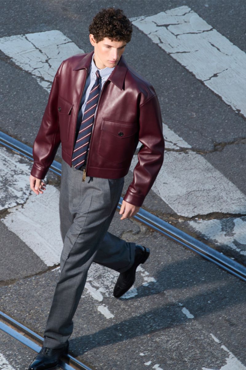 Fashionable man in leather jacket strides across zebra crossing with tramlines, styled in grey trousers and glossy black shoes.