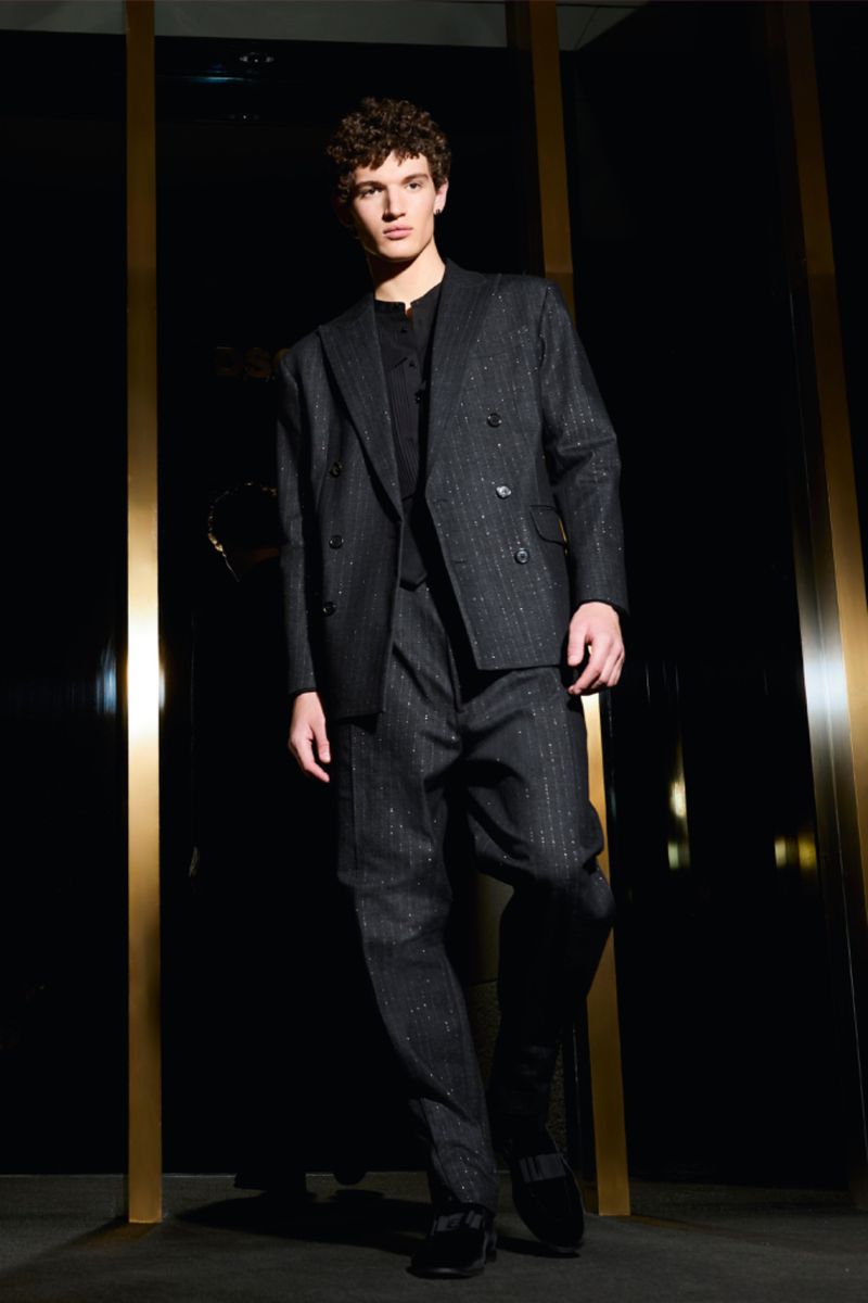 A fashionable adult male stands indoors wearing a dark grey striped double-breasted suit with dark shoes and a black background.