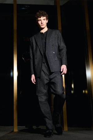 A fashionable adult male stands indoors wearing a dark grey striped double-breasted suit with dark shoes and a black background.