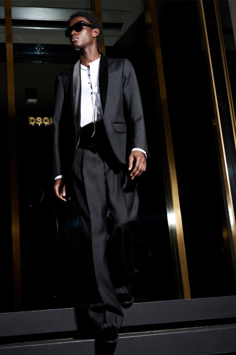 Man in black tuxedo with sunglasses standing on steps in front of a dark building with gold accents, fashion editorial.