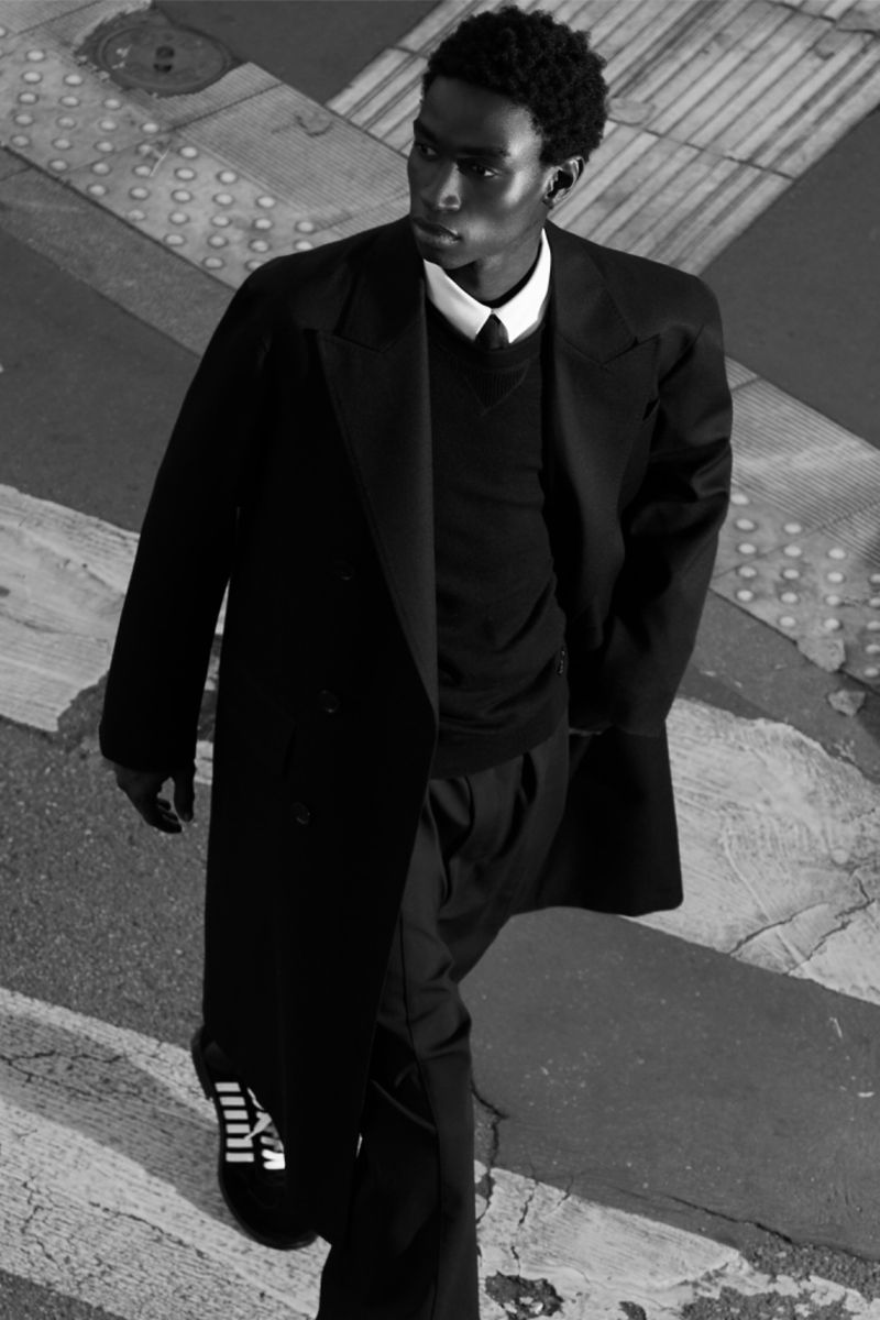 A stylish Black male in a long black coat walks across a crosswalk in a monochromatic fashion editorial style shot.