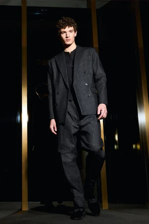 Model wearing a dark double-breasted suit standing indoors between vertical illuminated pillars.