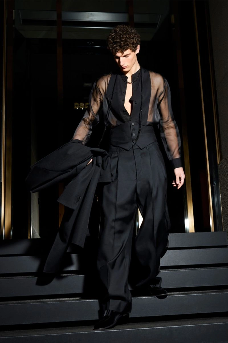 Male model in a black sheer shirt and trousers walking down dark steps holding a jacket