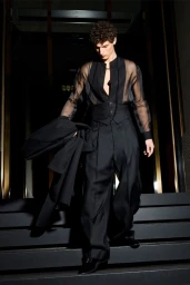 Male model in a black sheer shirt and trousers walking down dark steps holding a jacket