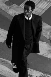 Black man wearing a dark coat and trousers walking across a paved street.