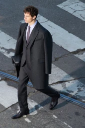 Model wearing a dark grey suit coat over trousers with a tie, walking across a paved street.