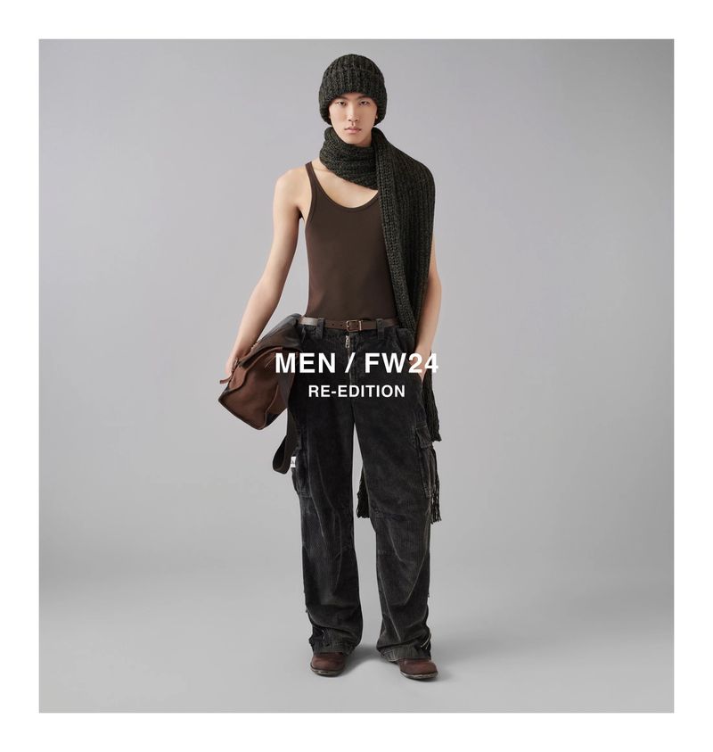 Model wearing a brown tank top, dark trousers, beanie, and knitted scarf while holding a brown bag against a grey seamless backdrop