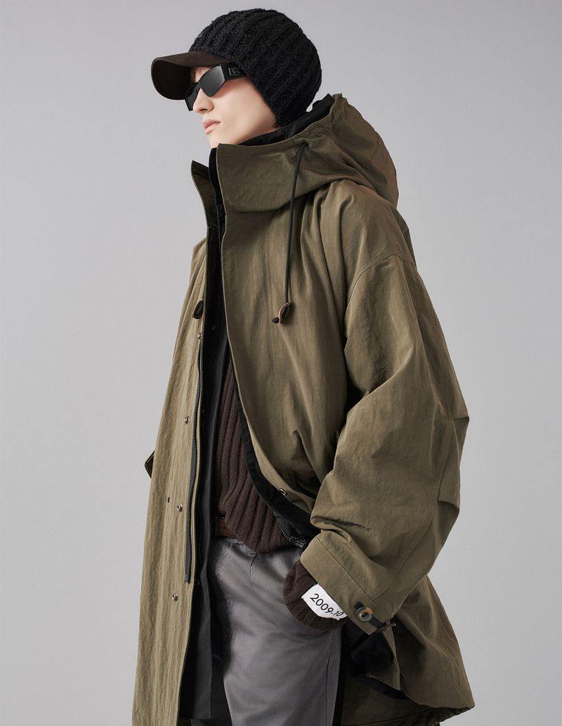 Man in olive green hooded trench coat with knit cap and sunglasses stands in a neutral studio. Fashion editorial with neutral colors.