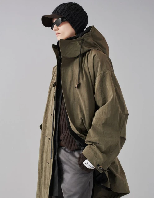 Man in olive green hooded trench coat with knit cap and sunglasses stands in a neutral studio. Fashion editorial with neutral colors.