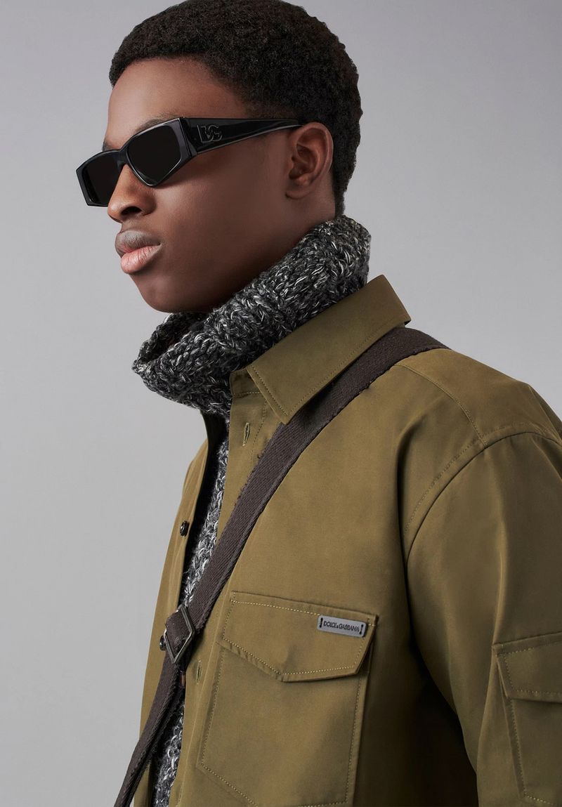 Studio shot of a black male model wearing a green collared shirt, grey sweater, and black sunglasses with a brown crossbody bag.