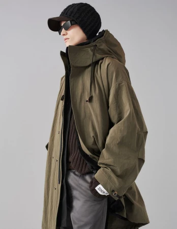 Model wearing an olive green hooded parka coat, black beanie, sunglasses, and grey trousers against a grey seamless background