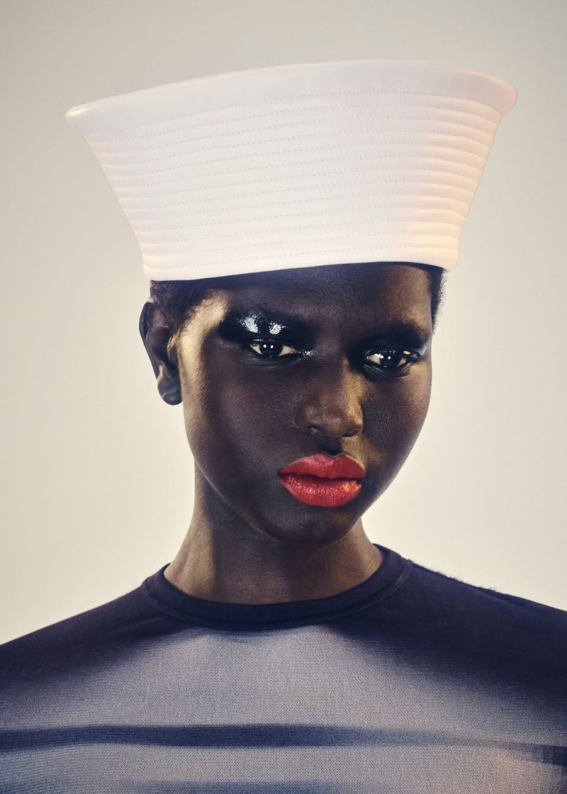 Close-up shot of a person with dark skin wearing a white sailor hat and red lipstick, gazing forward against a light background.