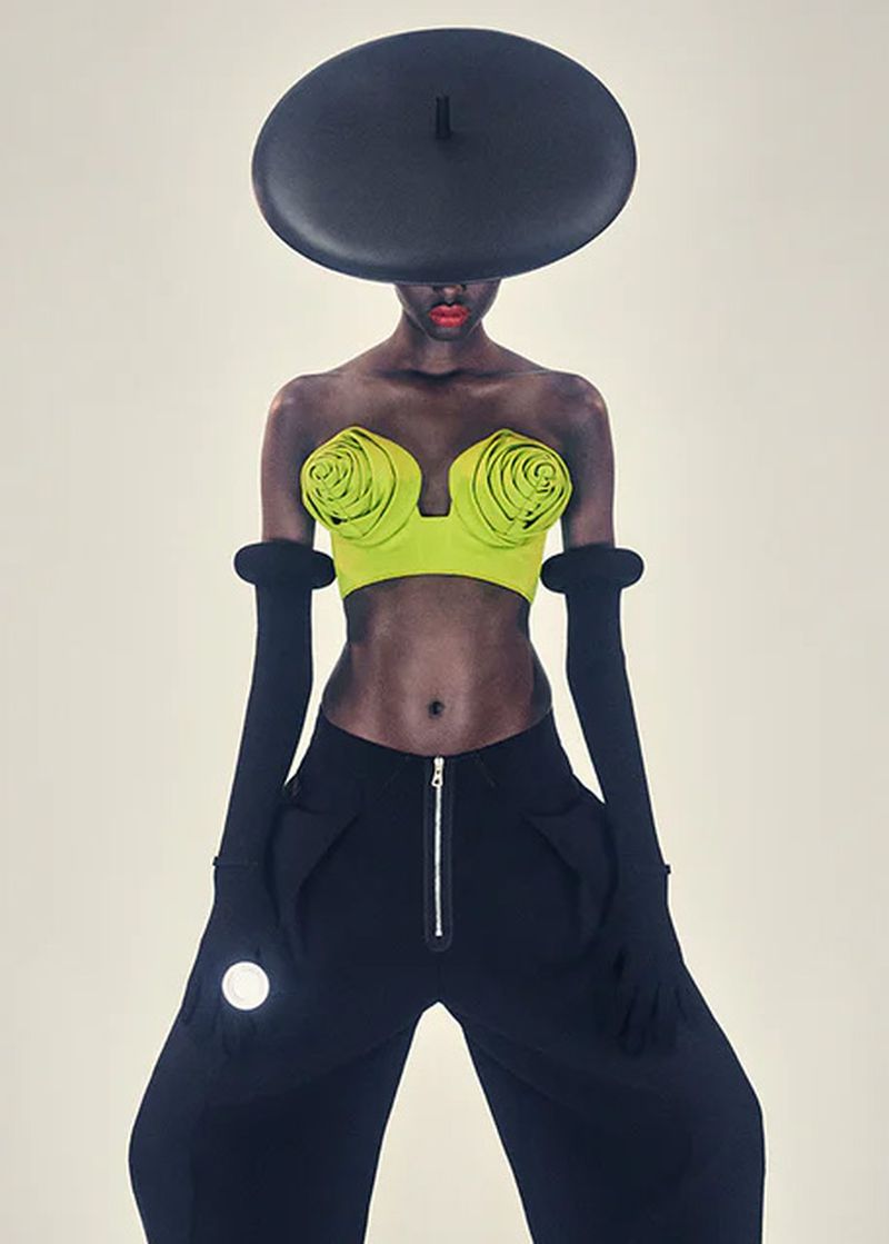 Fashionable woman with dark skin poses in modern clothing, including a lime top, black wide-leg pants, black gloves, and a large hat.