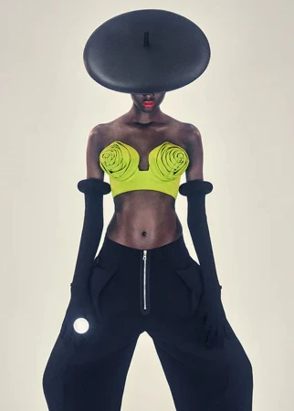 Fashionable woman with dark skin poses in modern clothing, including a lime top, black wide-leg pants, black gloves, and a large hat.
