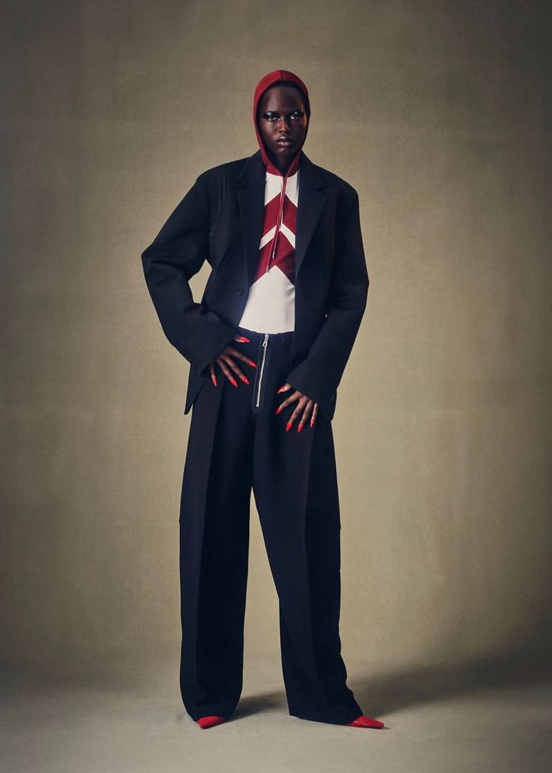 Full-length fashion portrait of an adult woman with dark skin wearing a black suit with a red hooded top and long red fingernails.