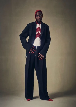 Full-length fashion portrait of an adult woman with dark skin wearing a black suit with a red hooded top and long red fingernails.