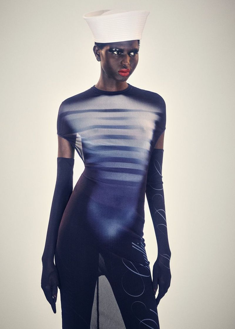 Woman with dark skin poses in a blue and white dress with long gloves and a white hat against a light background in a fashion studio shot.