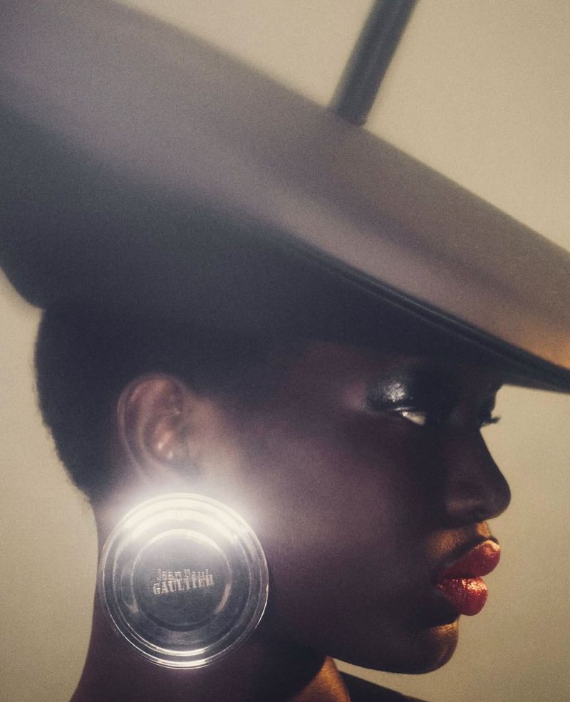 A dramatic fashion shot featuring a Black female model wearing a black hat and a large circular earring with the name of the designer.