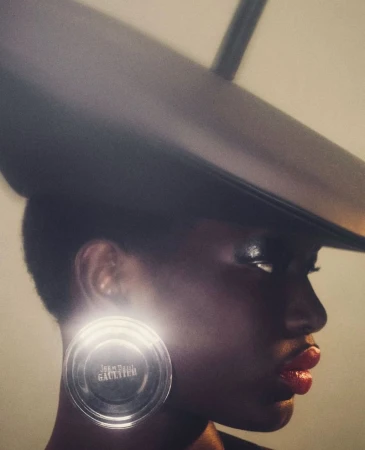 A dramatic fashion shot featuring a Black female model wearing a black hat and a large circular earring with the name of the designer.