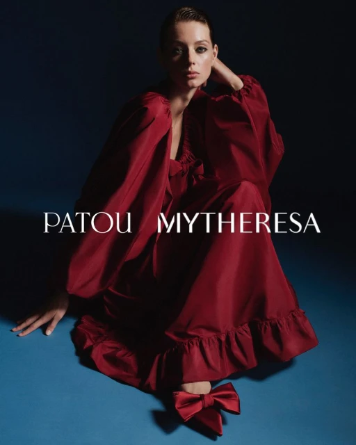 A poised fashion model in a flowing red dress and matching shoes, seated against a two-tone backdrop, exudes elegant style.