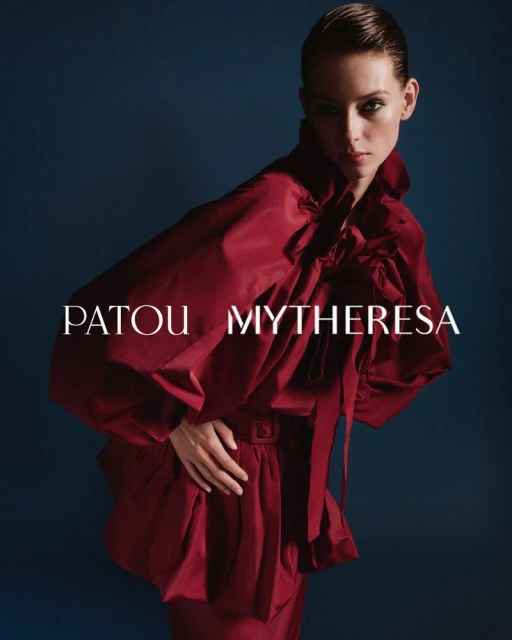 Fashion model poses in a draped, oversized, ruby-red Patou top with MYTHERESA text in a studio shot against a dark blue background.