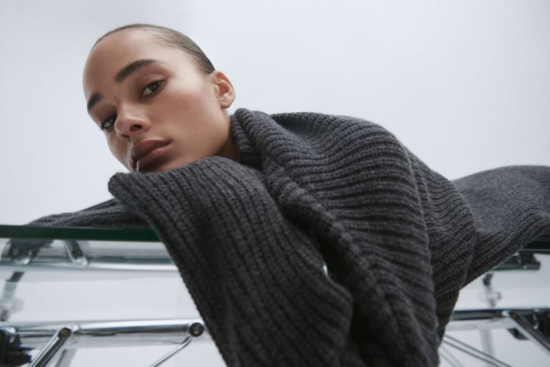 Woman with neutral expression poses in dark grey knit sweater on a glass table in a softly lit studio. Beauty and fashion portrait.