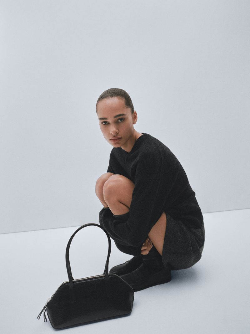 Model squatting in a black sweater with a structured black handbag placed on a white seamless studio floor.