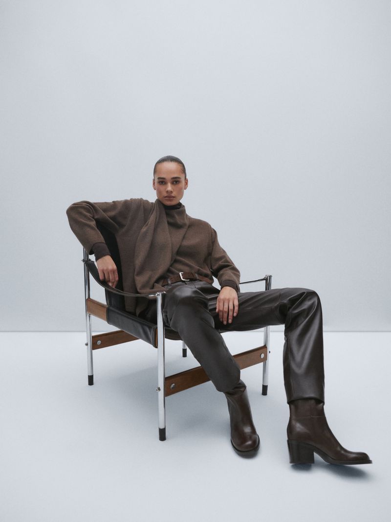 Fashion shot of a woman in brown sweater, leather pants, and ankle boots sitting in a mid-century modern chair on a white background.