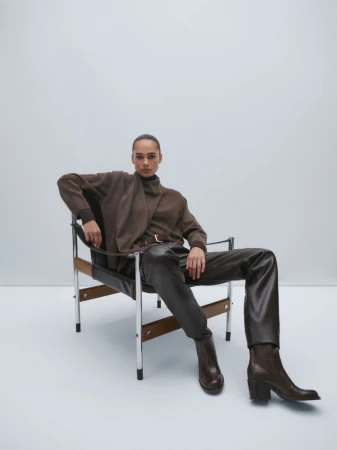 Fashion shot of a woman in brown sweater, leather pants, and ankle boots sitting in a mid-century modern chair on a white background.
