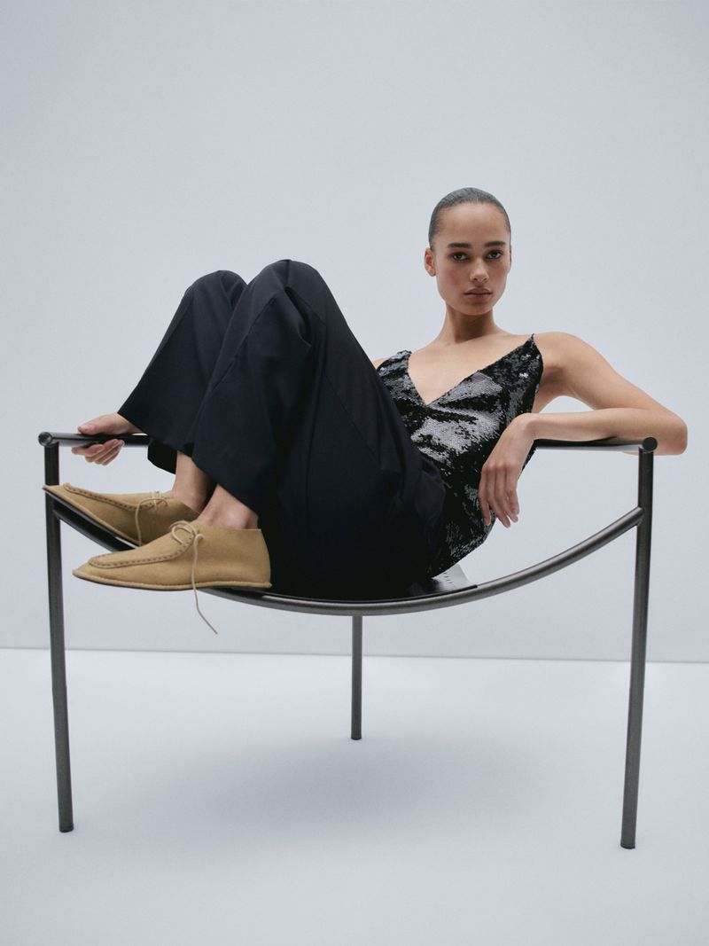 Model wearing dark trousers and camel shoes seated in a modern metal chair against a plain white studio background.