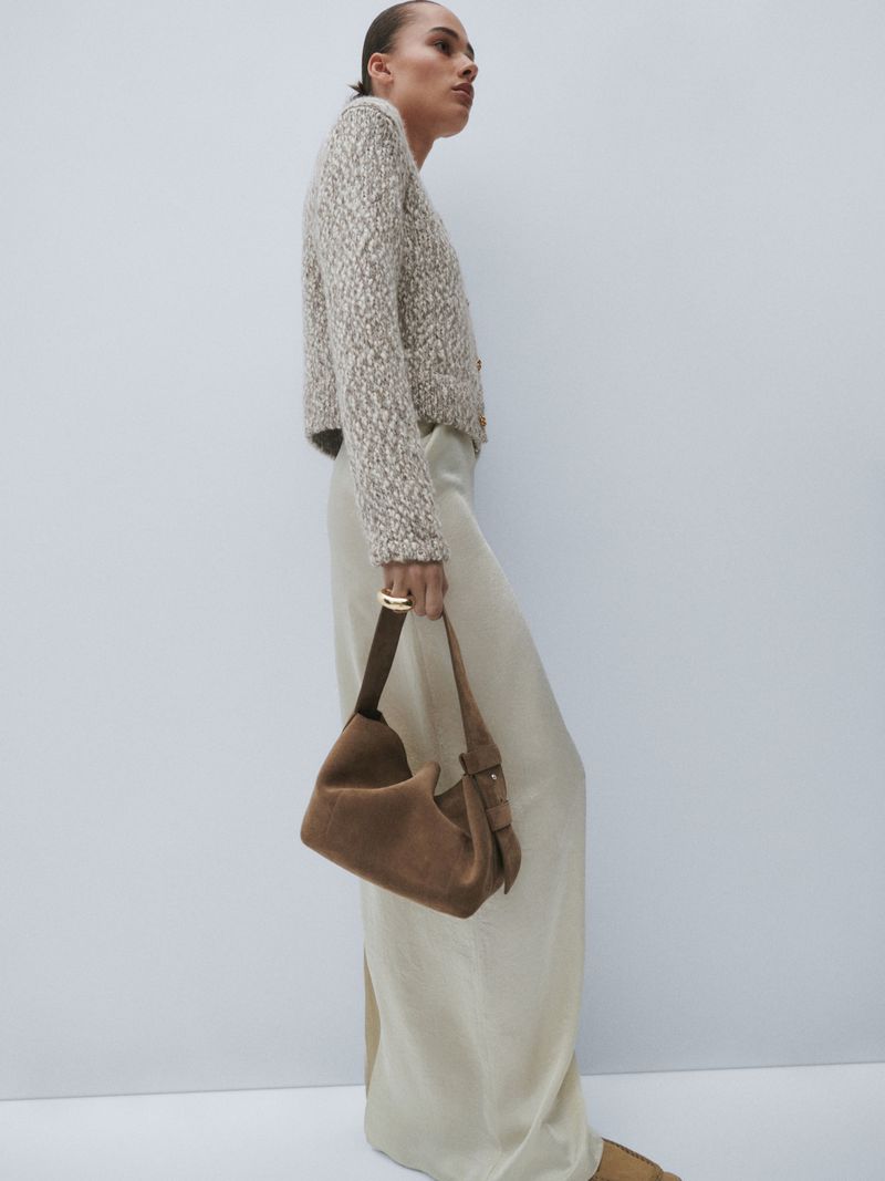 An adult female fashion model poses in a studio wearing a fuzzy cream sweater, wide leg satin trousers, and a suede shoulder bag.