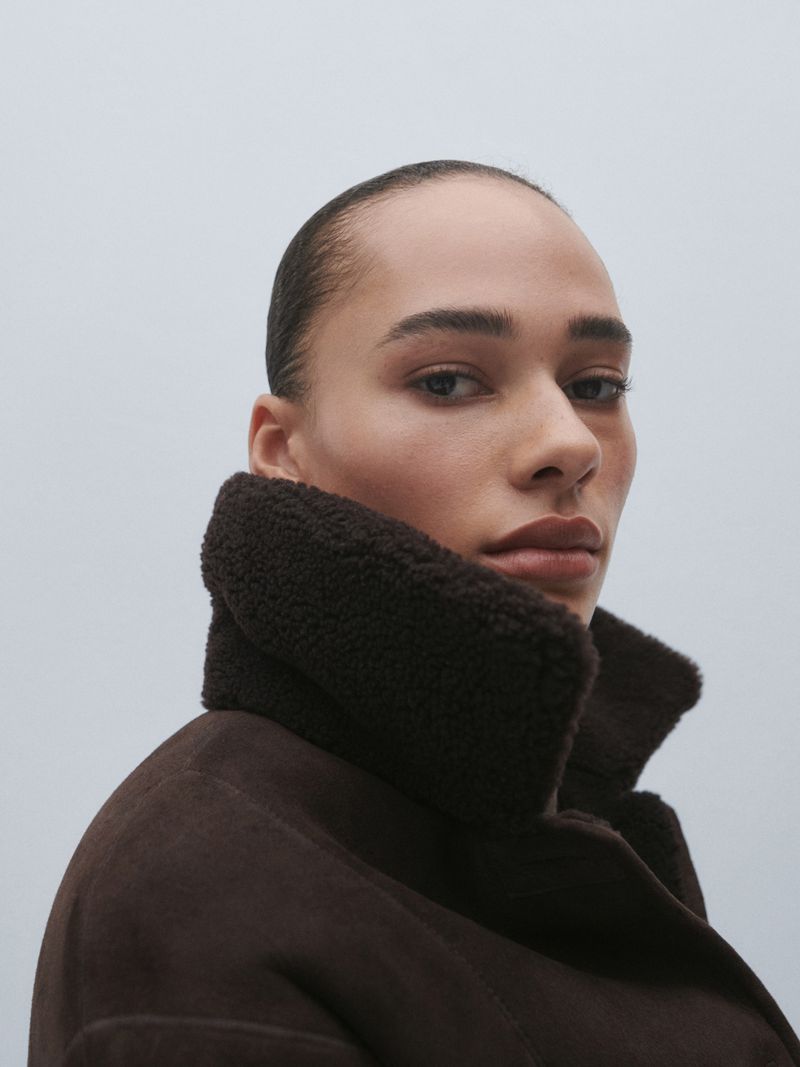 Close-up of a woman with slicked-back hair in a dark brown shearling coat. Low angle, natural light on a plain neutral background.