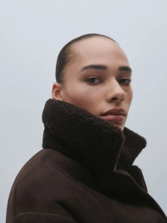 Close-up of a woman with slicked-back hair in a dark brown shearling coat. Low angle, natural light on a plain neutral background.