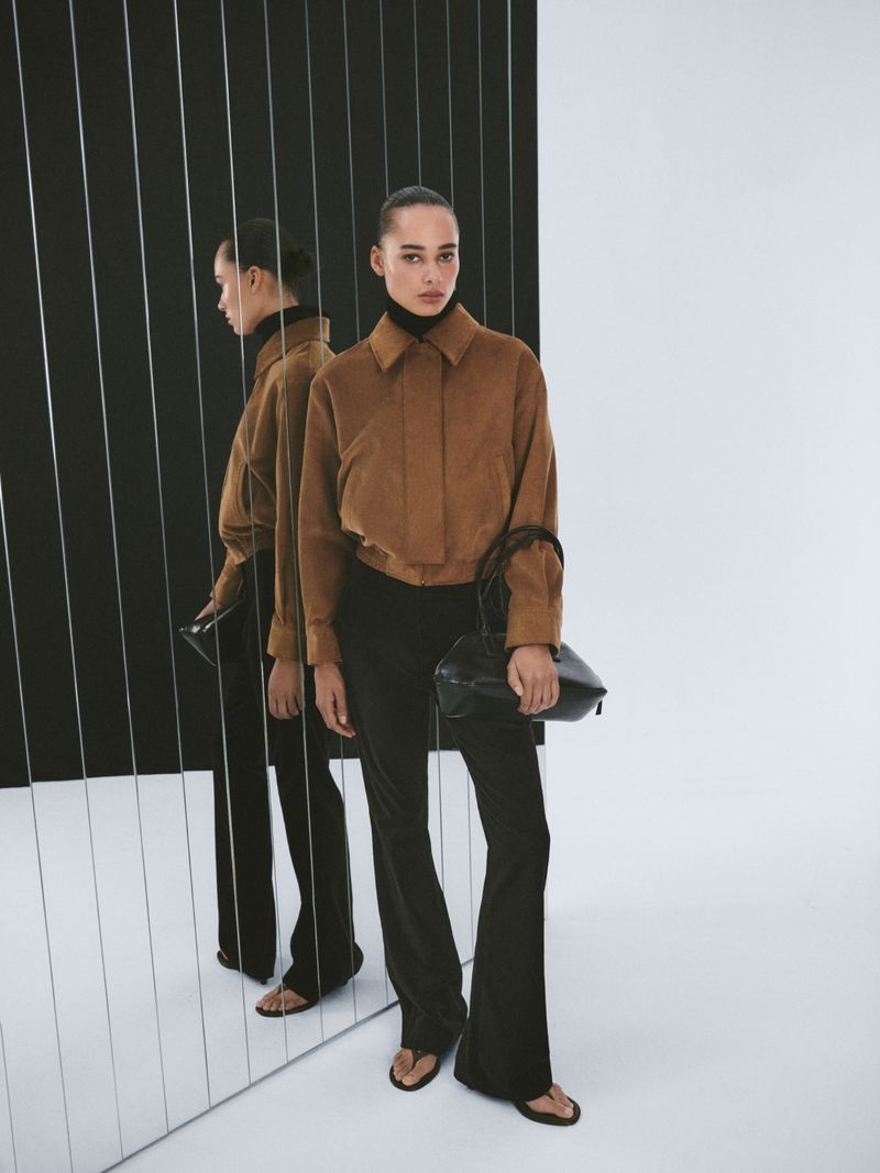 Brunette woman poses wearing a brown jacket, black pants, and a black bag with a black and white studio background and mirror reflection.