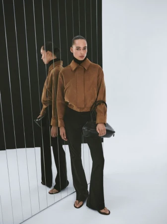 Brunette woman poses wearing a brown jacket, black pants, and a black bag with a black and white studio background and mirror reflection.