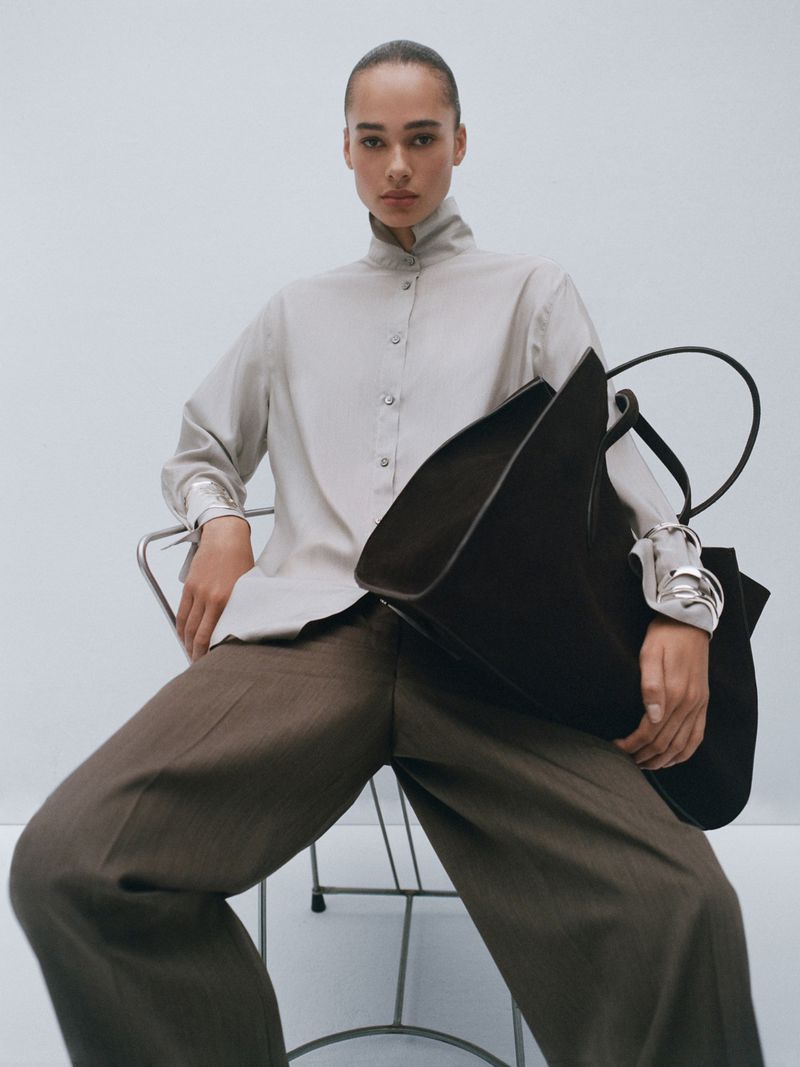 A fair-skinned female model is seated on a metal chair, wearing a light beige button-down shirt, brown slacks, and holding a black tote bag.