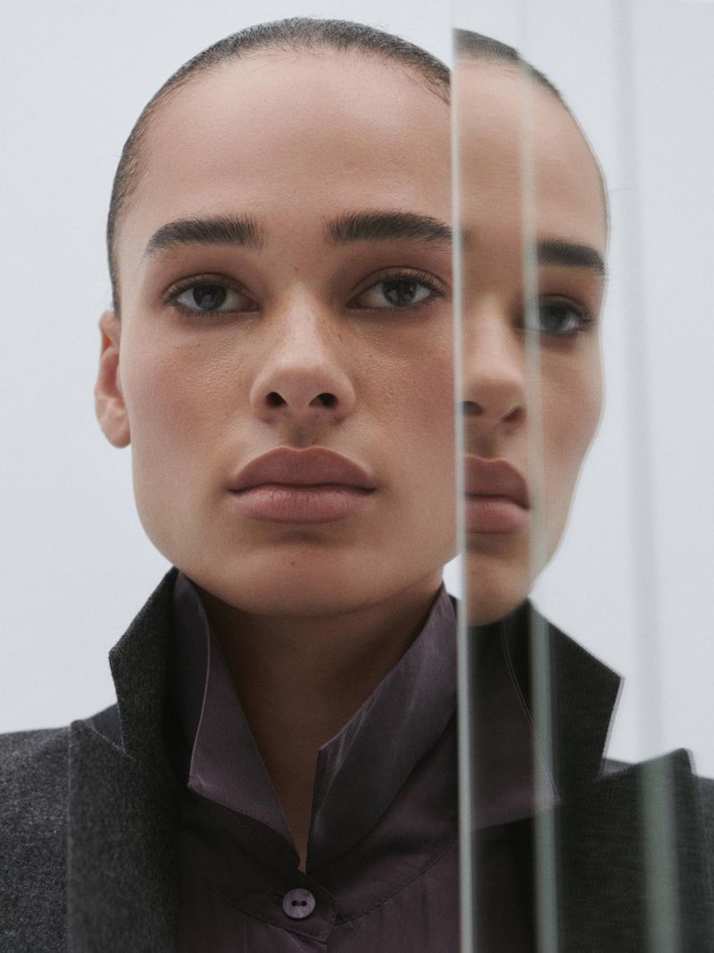 Close-up of an adult female model posing indoors in professional attire and makeup, reflected in glass against a white background.