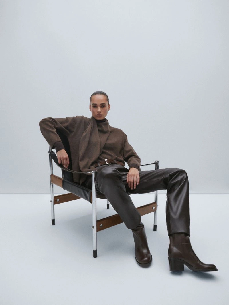 Model seated in a modern chrome and leather armchair wearing a brown sweater and dark leather trousers against a plain white background.