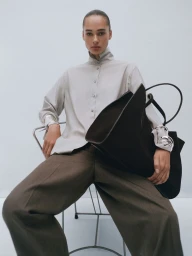 Model Annemary Aderibigbe seated on a chair holding a large dark tote bag against a plain white background for Massimo Dutti