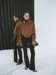 Model in brown jacket and black trousers holding a black handbag against a wall with vertical reflective panels.