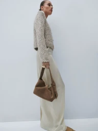Model standing against a plain white background wearing a textured cardigan and holding a brown suede handbag.