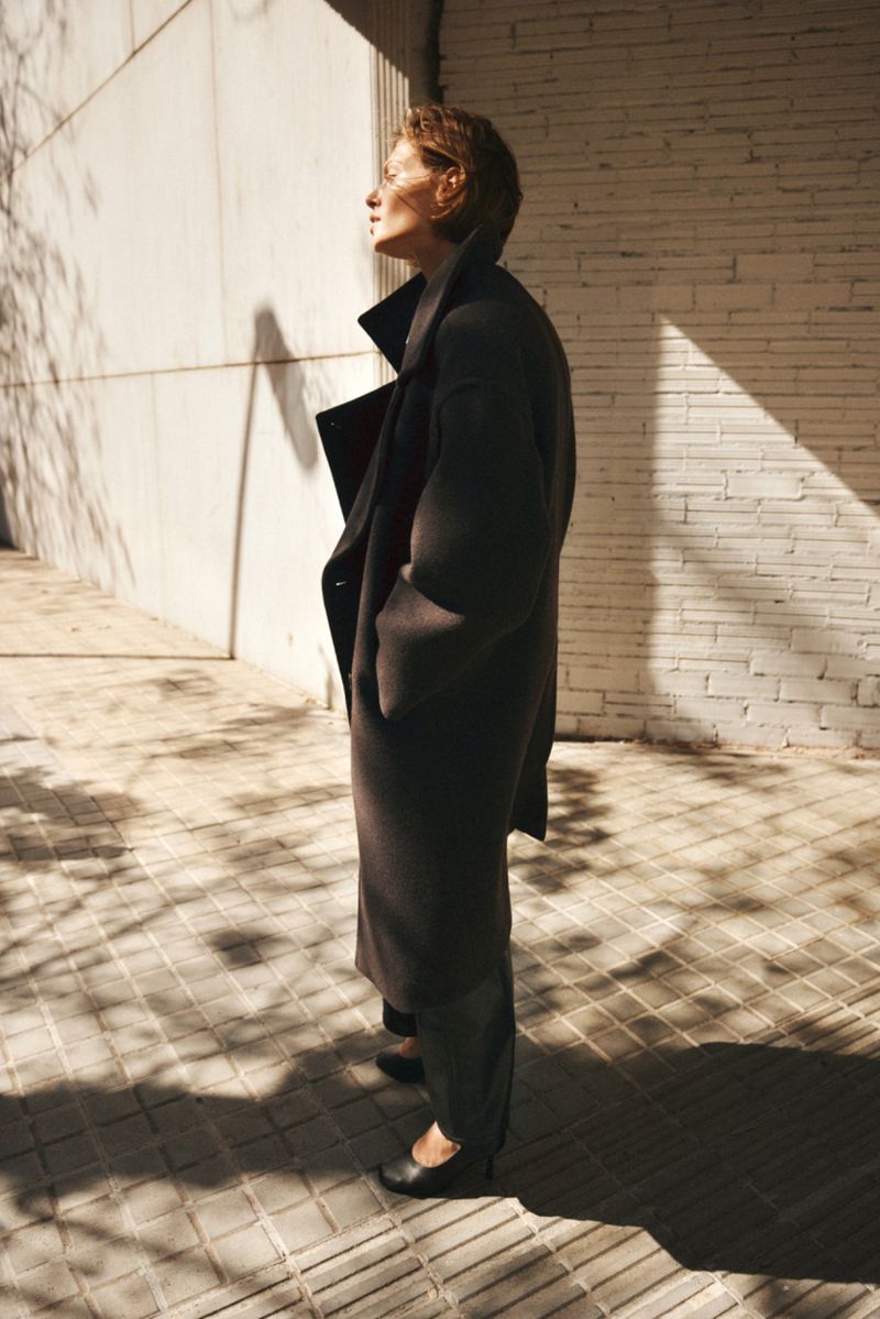 Model standing outdoors wearing a long dark coat and heels against a white brick wall in bright sunlight