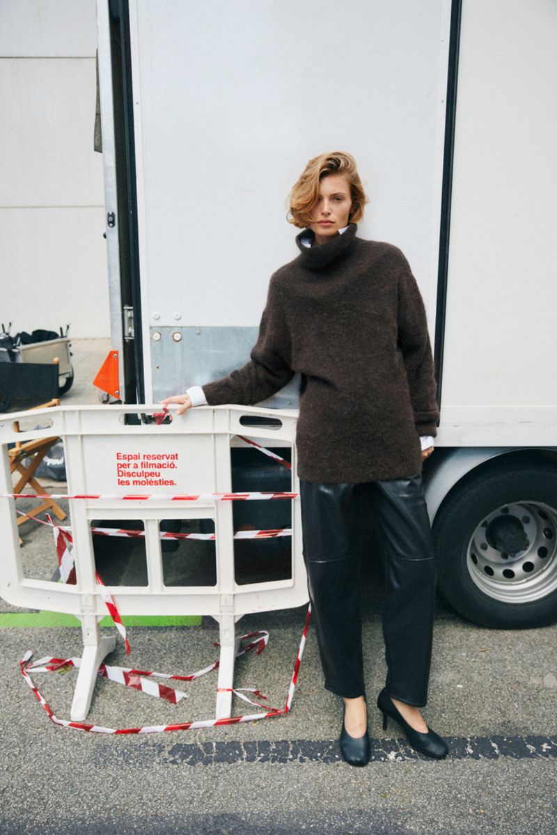Woman in oversized dark brown sweater dress and black heels poses next to a white truck and barricade in urban setting.