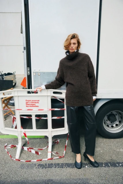 Woman in oversized dark brown sweater dress and black heels poses next to a white truck and barricade in urban setting.