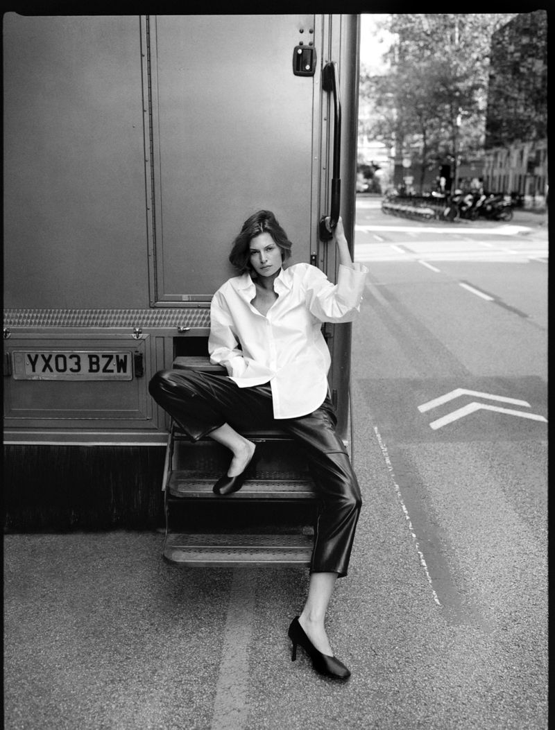 A striking black and white fashion shot of a woman in leather pants and a white shirt sitting on bus steps on a city street.