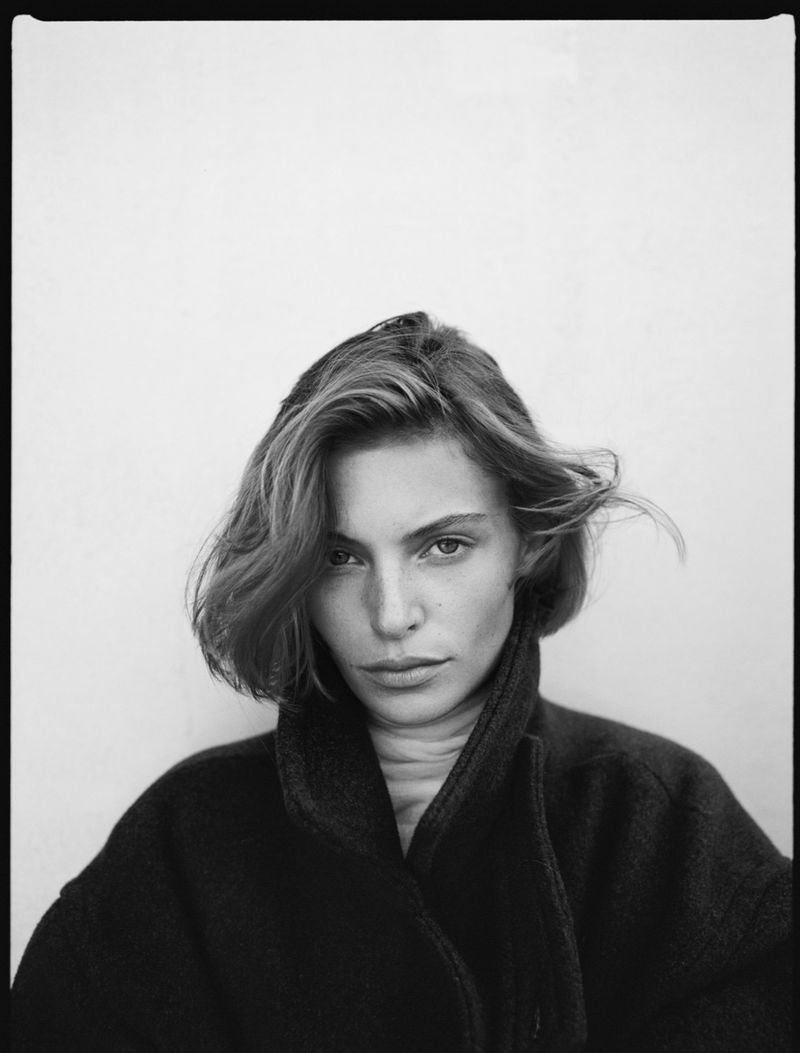 Close-up portrait of a woman with short dark hair wearing a high-collared dark coat against a plain background.