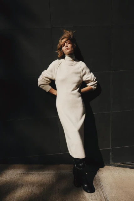 Woman in cream-colored dress with hands on hips leans against a dark wall, with sunlight casting shadows on the wall and floor.