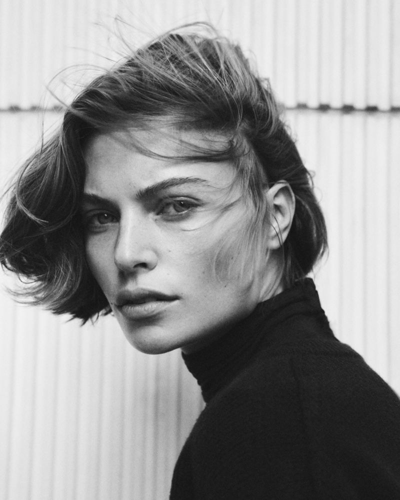 Close-up portrait of a woman with short hair wearing a black turtleneck against a textured background